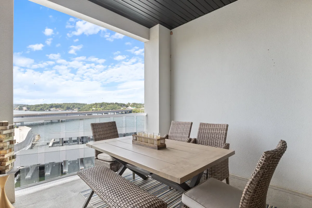 A spacious outdoor balcony featuring a dining table with woven chairs, set against a stunning panoramic lake view and open sky. The arrangement invites guests to dine al fresco while soaking up the tranquil water scenery. The setting suggests both morning and evening enjoyment, with ample space for groups or couples.