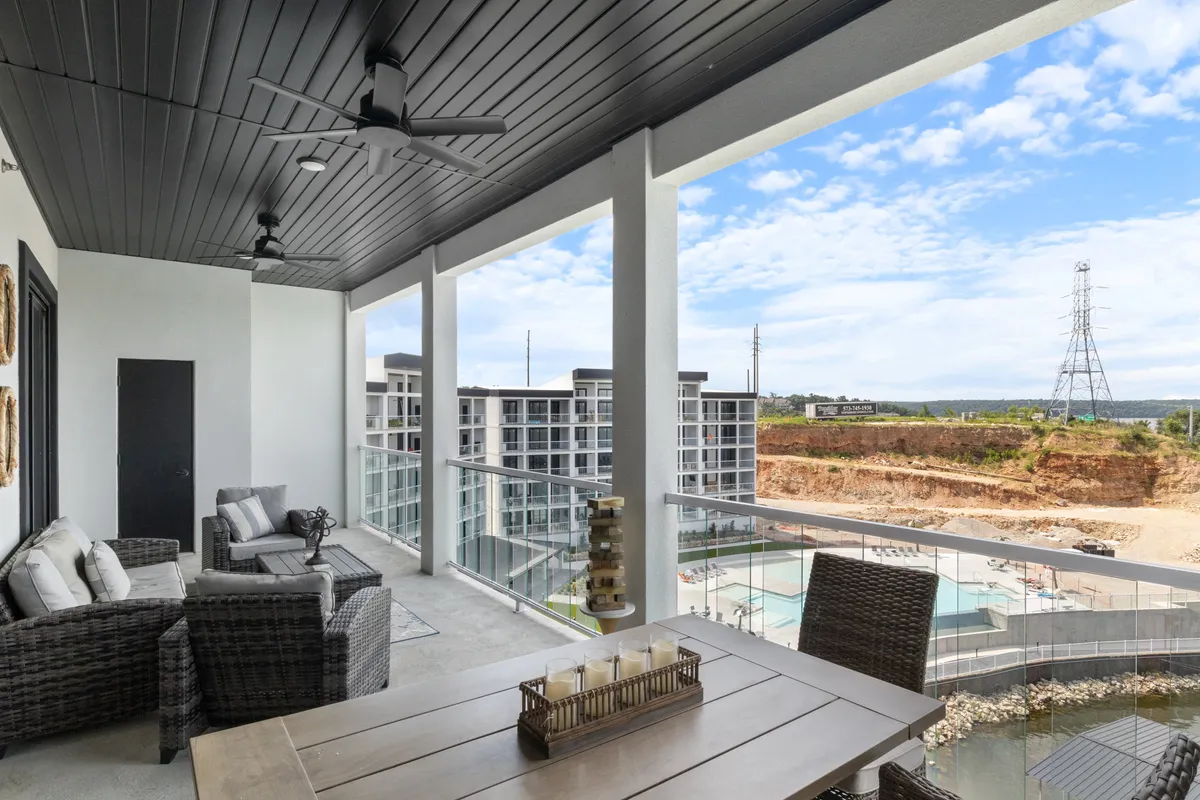 A spacious balcony with a comfortable outdoor seating area and dining table overlooks a sparkling pool area, surrounded by lush landscape. The setting is ideal for gatherings, meals, and sunset views, with the pool and social spaces visible just beyond the railing.