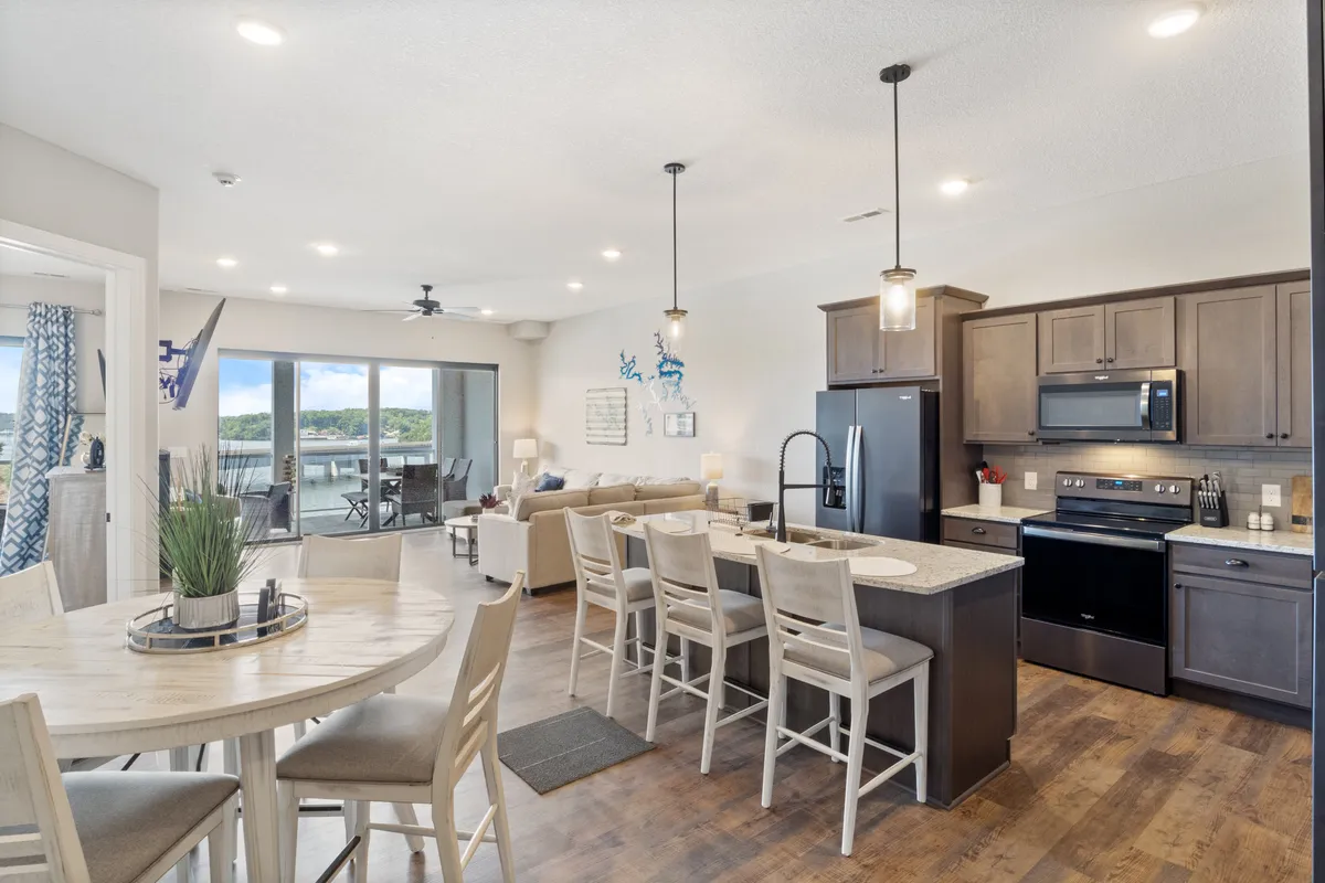 A spacious, open-plan area featuring a kitchen island, dining table, and a comfortable living area. Large windows allow abundant natural light to fill the space, offering scenic views of the lake outside. The layout invites social gatherings and seamless movement between cooking, dining, and relaxing.