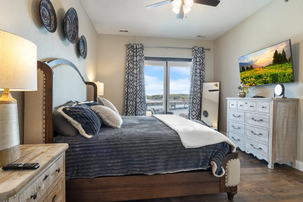 This image features a serene bedroom with a bed adorned with decorative pillows, a large window offering a view, a wall-mounted television, and a dresser. Natural light floods the space, creating a tranquil and airy atmosphere ideal for relaxation and a restful night's sleep.