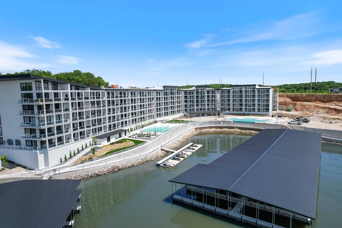 A panoramic exterior view showcasing the modern building complex with multiple swimming pools in the foreground and a marina filled with boats in the background. The water glistens under the daylight, and there are sunbathers and families enjoying the outdoor amenities, creating a lively and upscale atmosphere.